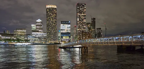 This urban landscape photograph captures Canary Wharf Night Pier 2 in London, United Kingdom, during the evening in autumn. The image shows modern high-rise buildings of Canary Wharf with illuminated windows reflecting on the waters of the River Thames. Notable landmarks such as One Canada Square and the distinctive cylindrical Newfoundland Tower are visible among the city’s skyline. The well-lit pier structure extends out onto the River Thames, demonstrating the urban architecture that characterizes this financial district in London.